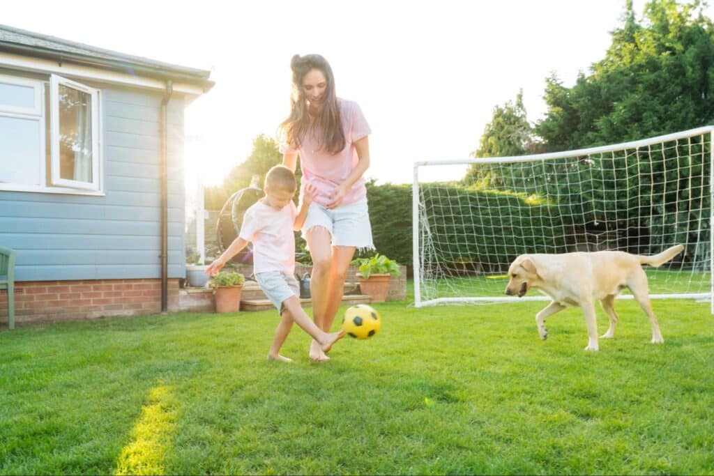 Family playing soccer outdoors with a dog in a sunny backyard.