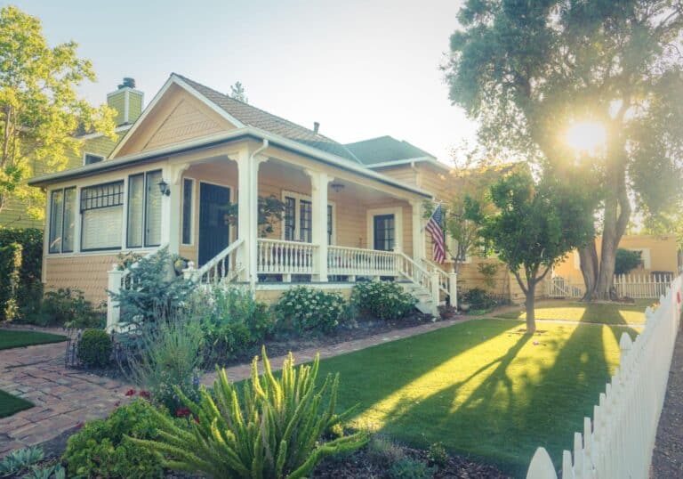 Charming yellow house with porch, beautiful landscaping, and sunlight filtering through trees.